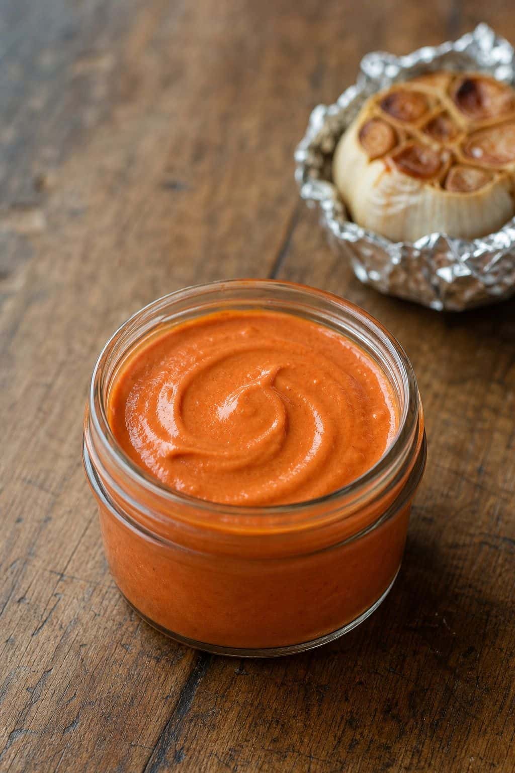 Gochujang Aioli Recipe (Spicy Korean Garlic Mayo) Creamy gochujang aioli in a glass jar with roasted garlic in the background