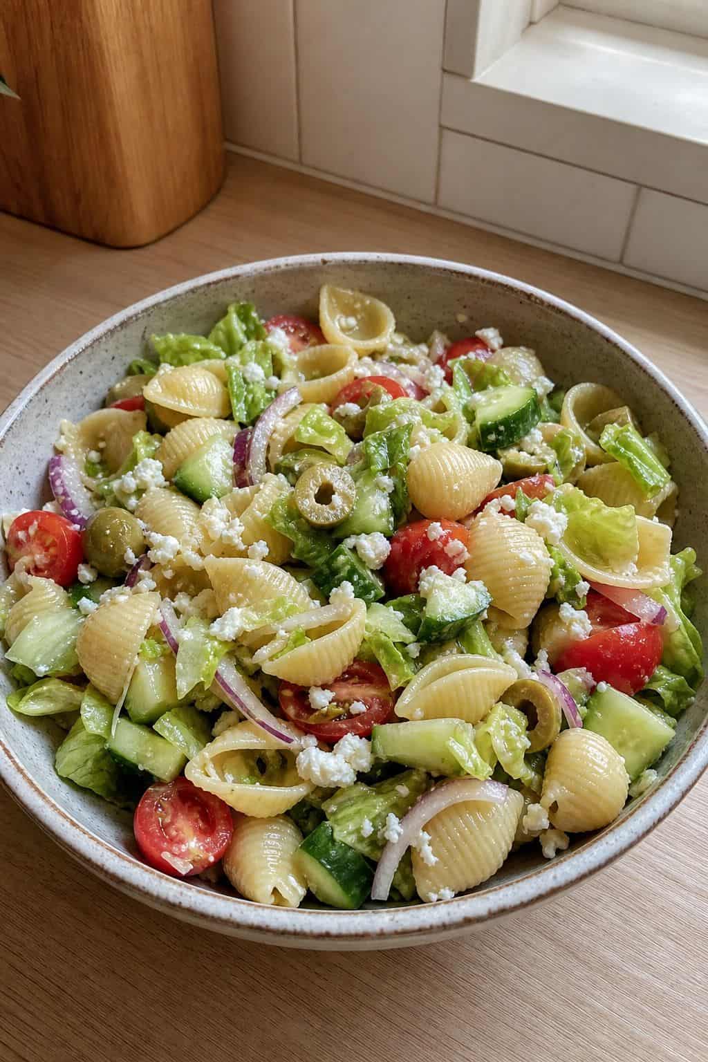 Greek pasta salad with shell pasta, feta, tomatoes, cucumber, red onion, lettuce, and green olives in a ceramic bowl.