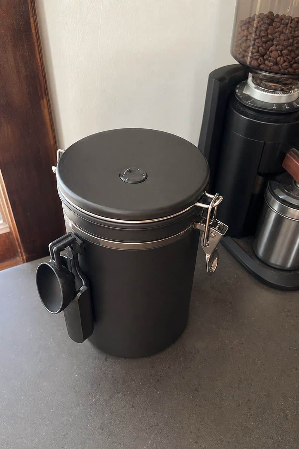 An opaque sealed canister near the grinder protects coffee and keeps an attached scoop easy to reach.
