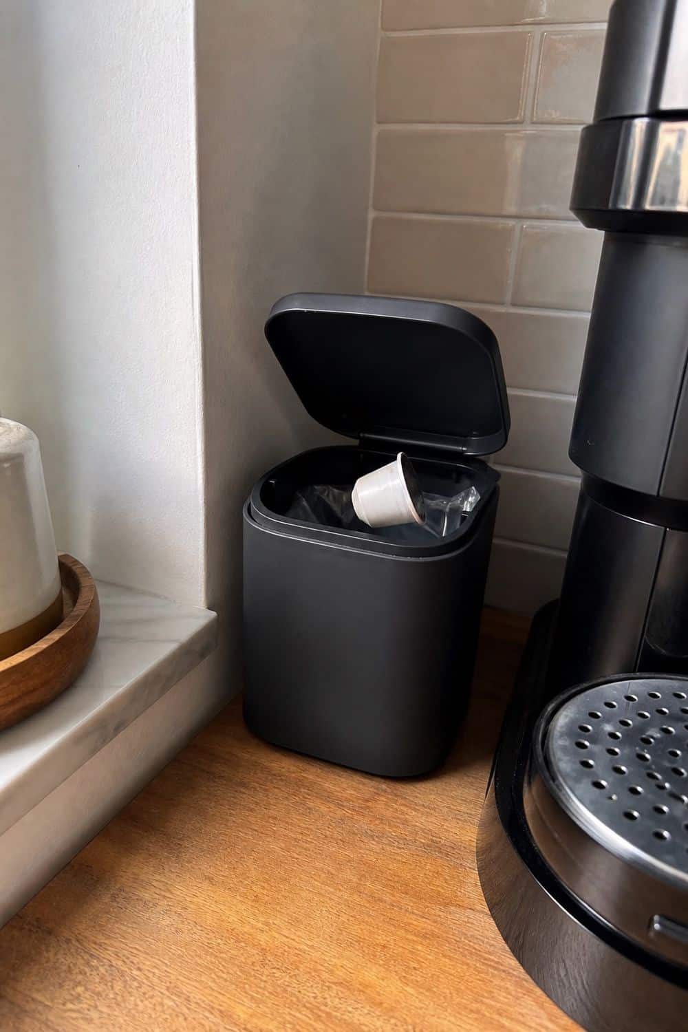 A small lidded trash can with removable liner is tucked by the machine for pods and wrappers.